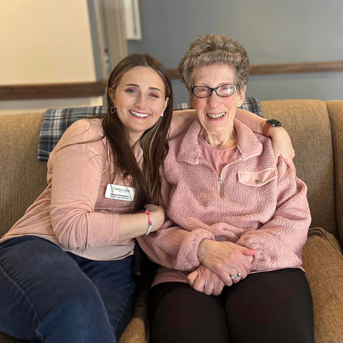 Senior resident and team member, both dressed in pink, sit together on a couch, smiling and enjoying a relaxed moment, showcasing the warmth and camaraderie in the community.