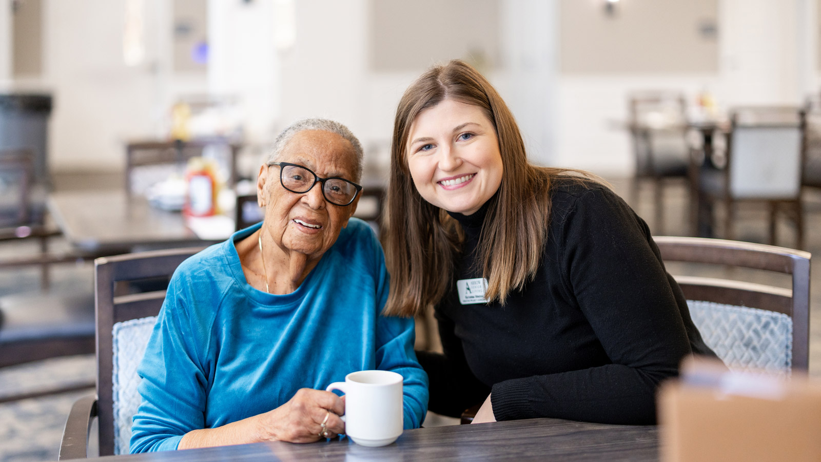 Smiling elderly woman with glasses and a blue shirt sitting at a table with a mug, alongside a younger woman in a black sweater, in a bright indoor setting.