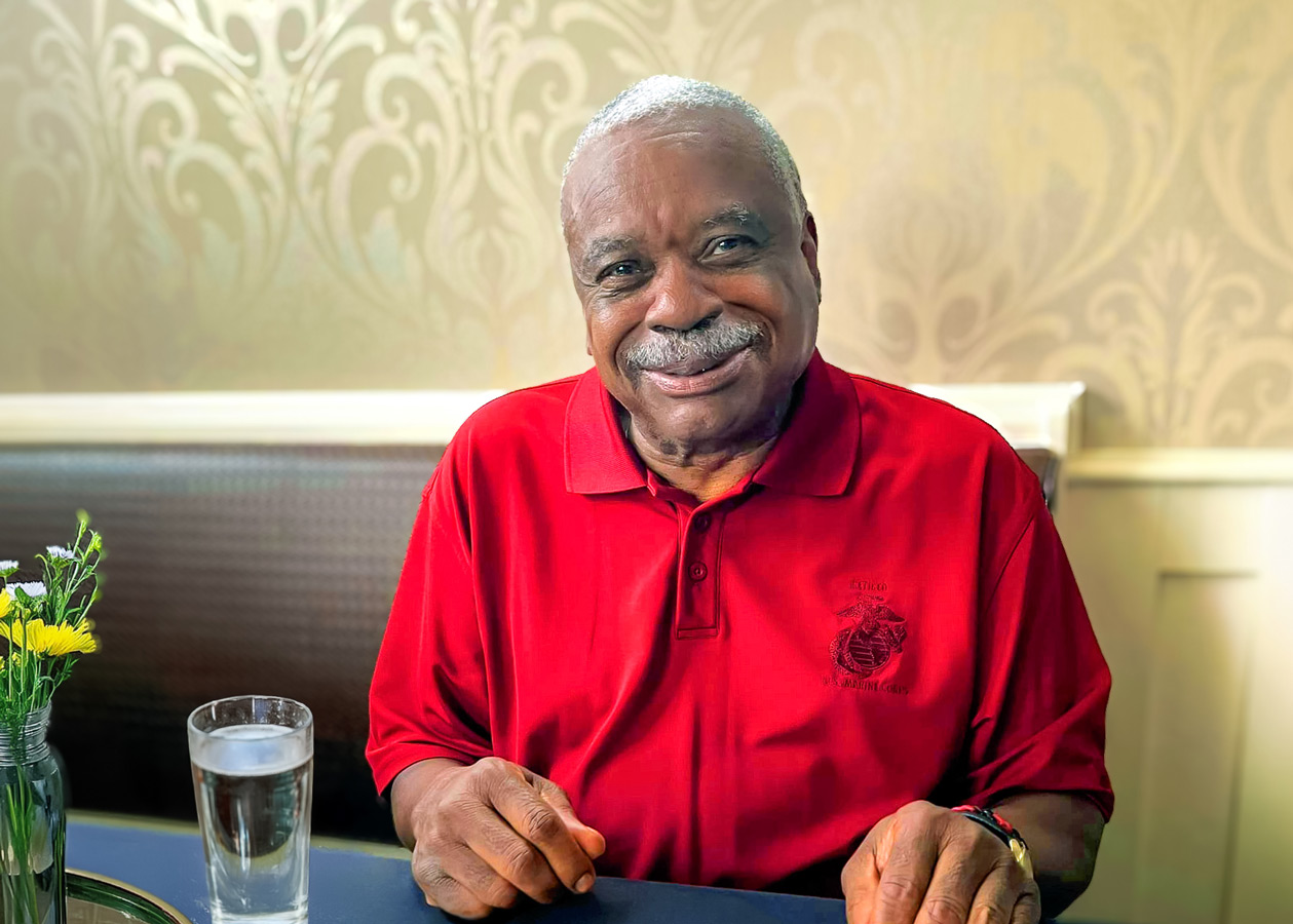Smiling senior resident in a red polo shirt with a U.S. Marine Corps logo, seated at a table against an elegant patterned wallpaper, radiating warmth and pride in community living.