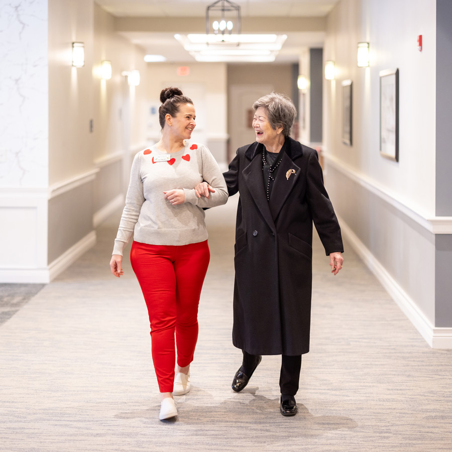 A senior resident enjoys a friendly walk with a team member through a brightly lit hallway. The team member wears a festive heart-themed sweater, sharing smiles and conversation.