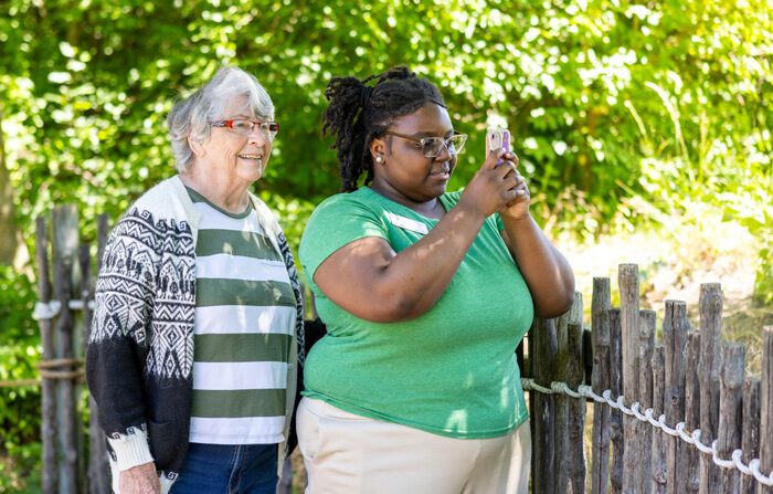 Two women enjoying a sunny day outdoors, with one taking a photo on her phone and the other smiling beside her, standing by a wooden fence surrounded by lush greenery.