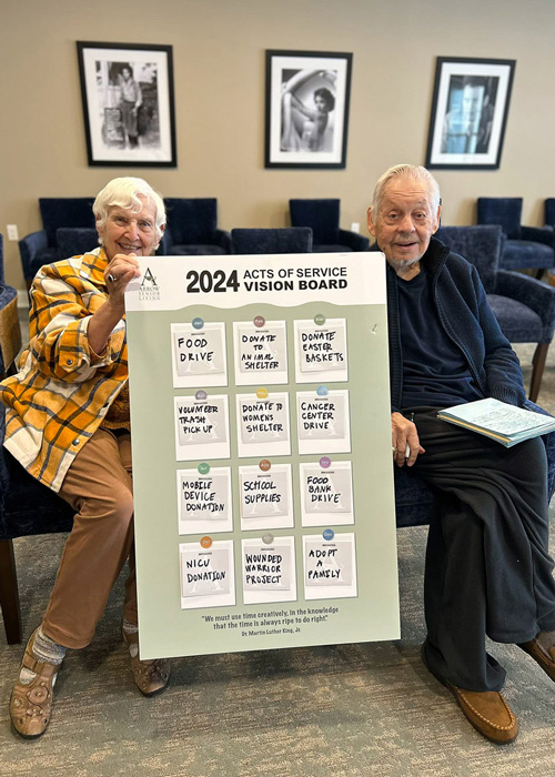 Two senior residents proudly display a large '2024 Acts of Service Vision Board' featuring various charitable activities, including food drives, donations, and volunteer work.