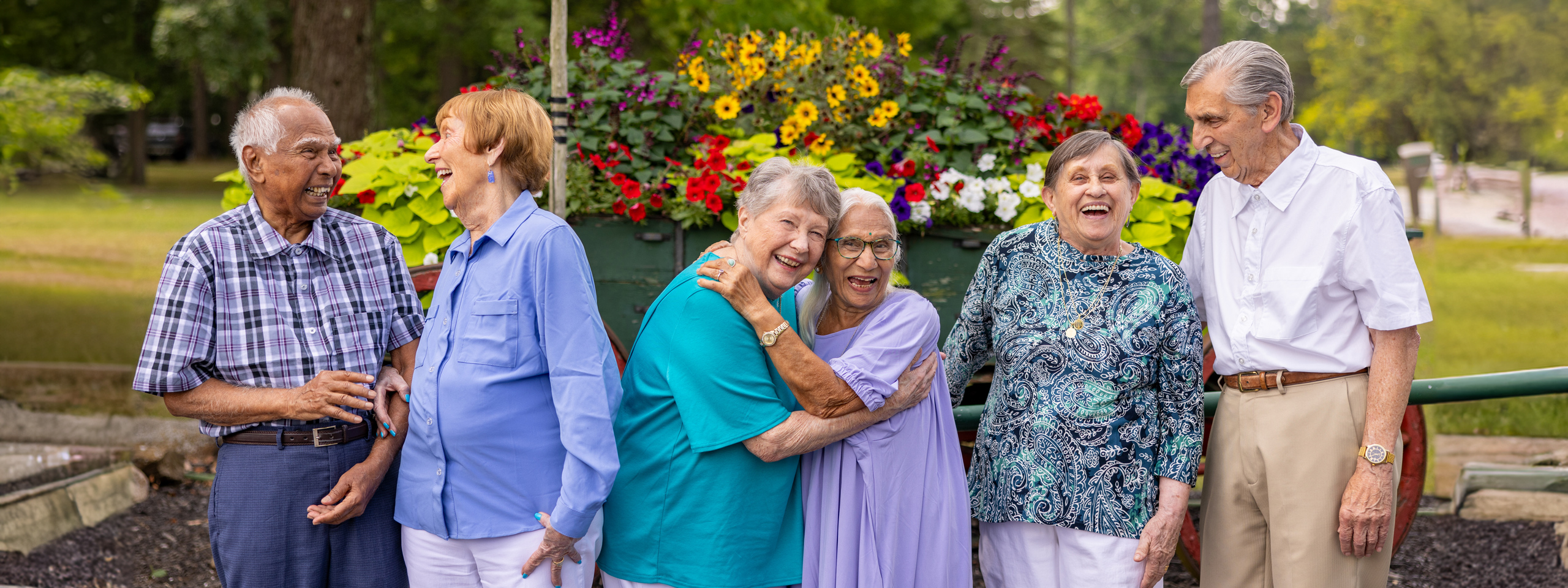 Group of six seniors laughing and enjoying each other's company outdoors, standing in front of vibrant flowers, showcasing joy, friendship, and the beauty of community living.