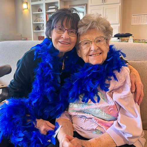 Two senior resident women sitting close together on a couch, both smiling and wearing a bright blue feather boa around their necks, enjoying a lively moment in a senior living community.