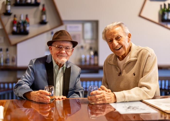 Two senior residents, one wearing a stylish hat and blazer, the other in a casual sweater, sit together at a bar, smiling warmly and enjoying drinks in a relaxed atmosphere.