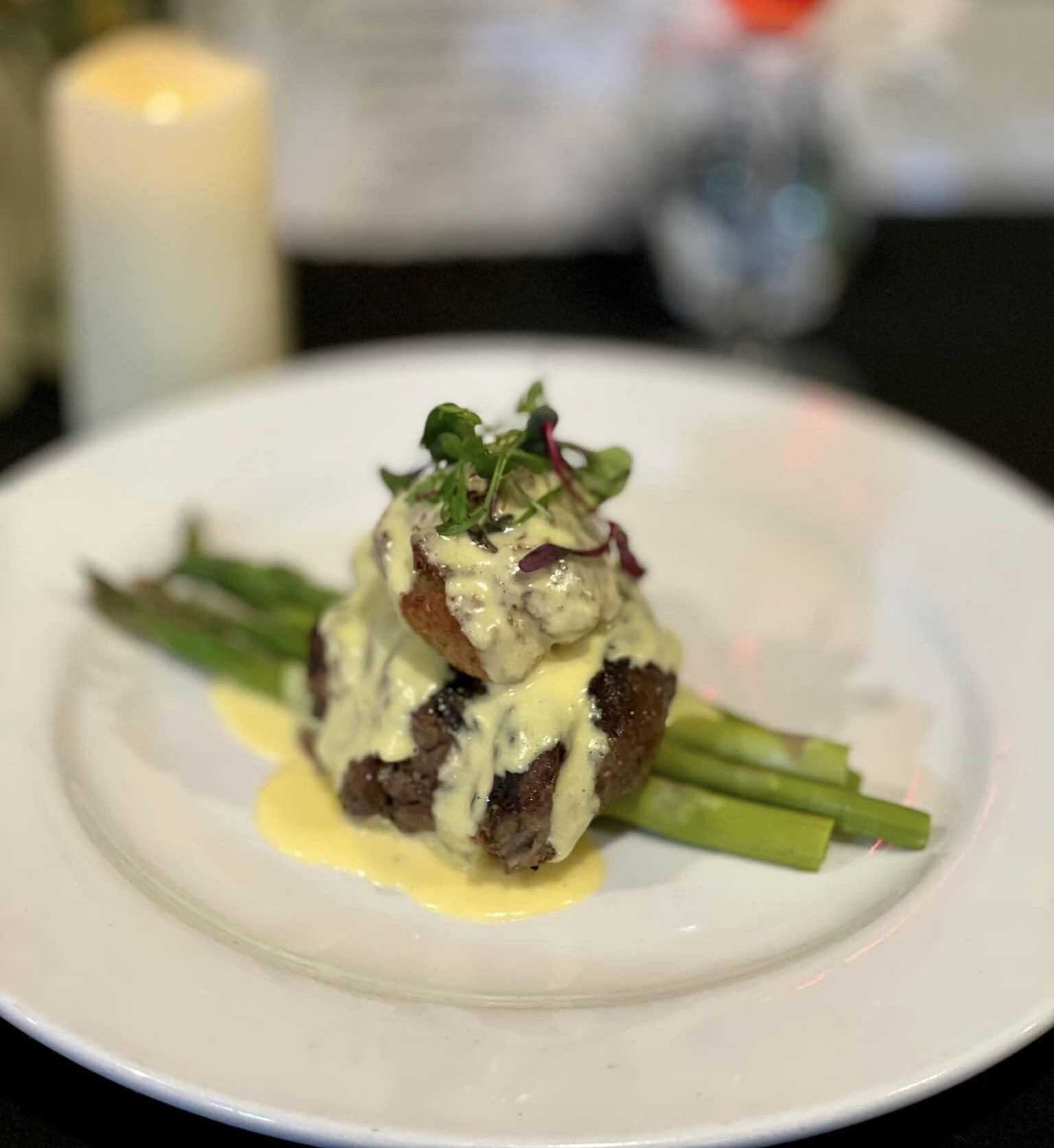 Plated gourmet steak with asparagus, topped with creamy hollandaise sauce and garnished with microgreens, set against a blurred background of a candle, flowers, and drink.