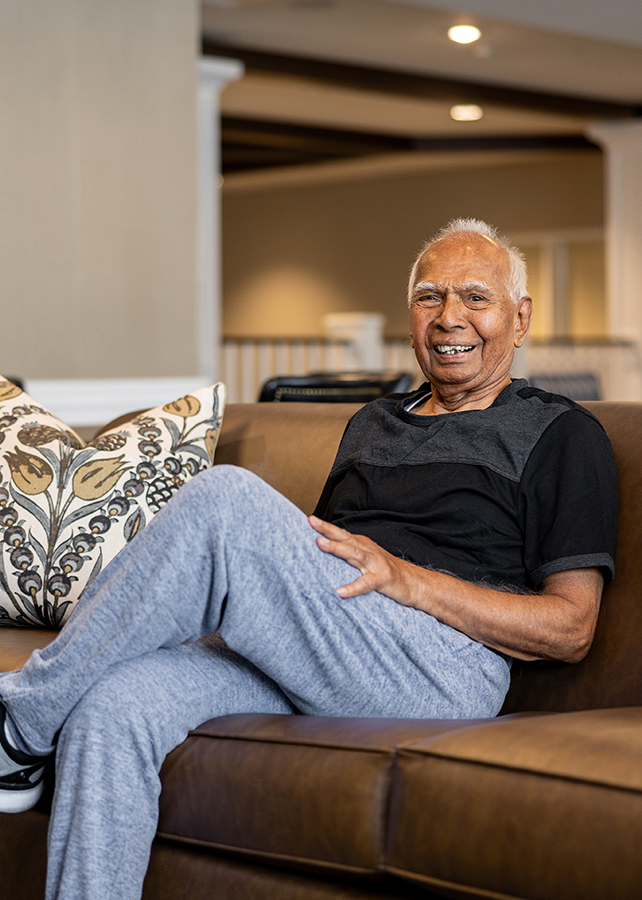 Senior resident relaxing on a brown couch, smiling in a cozy lounge area with patterned pillows, creating a welcoming and comfortable atmosphere.