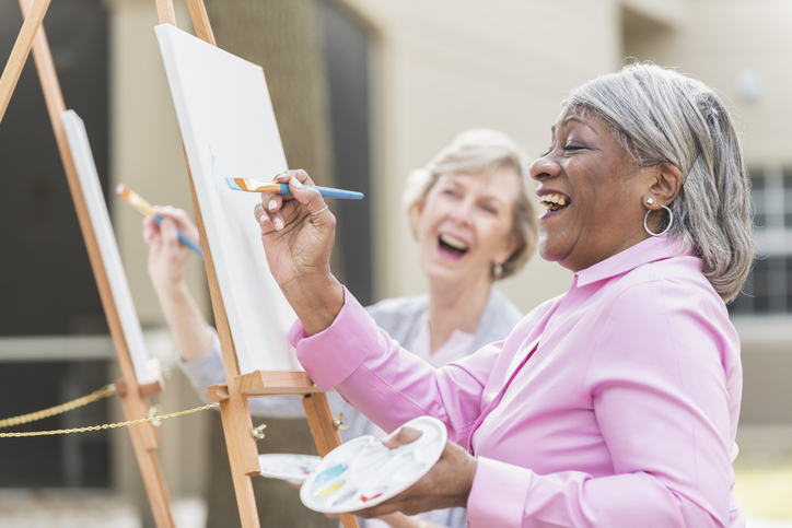 Two senior women joyfully painting on easels outdoors during an art class