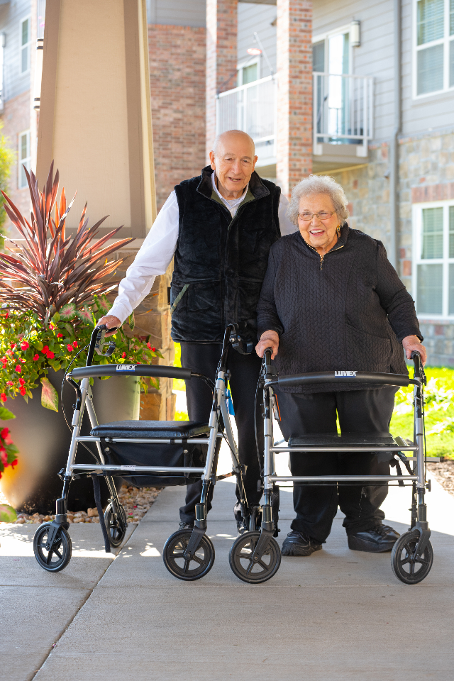 Two senior residents smile while walking side by side outdoors using walkers, enjoying fresh air on a landscaped walkway at a senior living community.