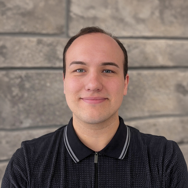 Tyler Dunbar, Senior Living Director at The Riverside Senior Living at Maumelle, in a professional headshot with short dark hair, a dark collared shirt, and a gray stone wall background.
