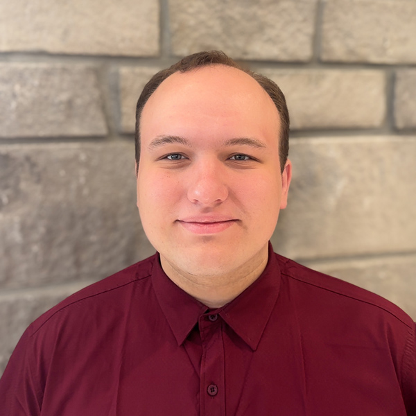 Tyler Dunbar, Senior Living Director at The Riverside Senior Living, smiling slightly while wearing a maroon button-up shirt, standing in front of a stone wall backdrop.