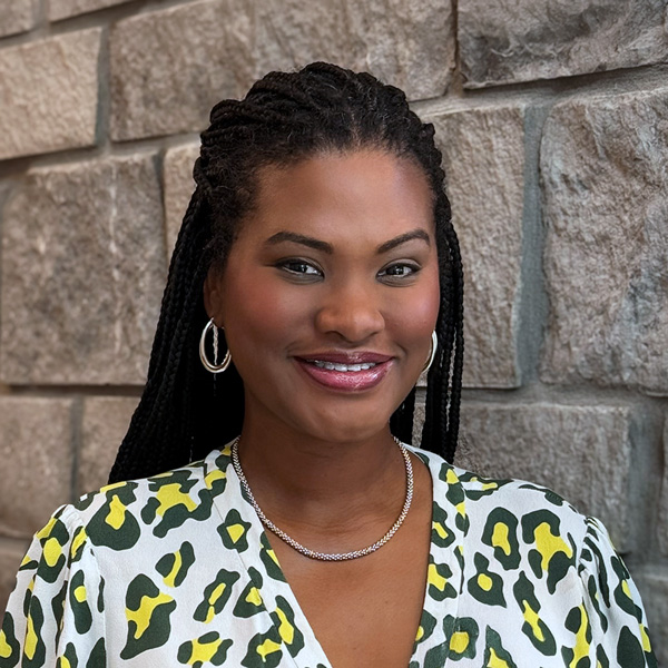 Michelle DuVall, Senior Living Coordinator at The Riverside Senior Living, smiling in a professional headshot with long braids, hoop earrings, a patterned blouse, and a gray stone wall background.